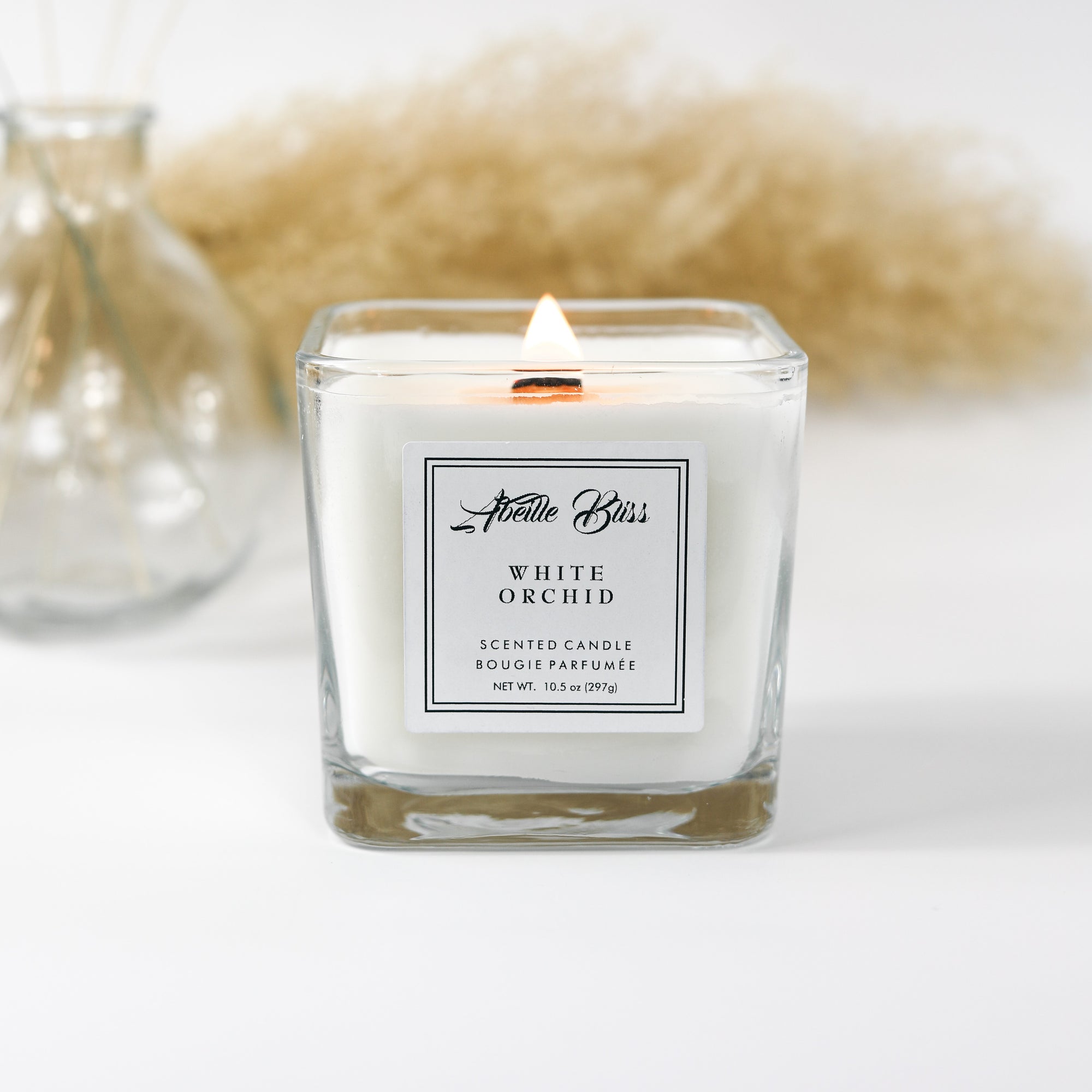 A white orchid candle with a clear glass container, featuring a label with the brand name 'Abeille Bliss' and the scent 'White Orchid'.