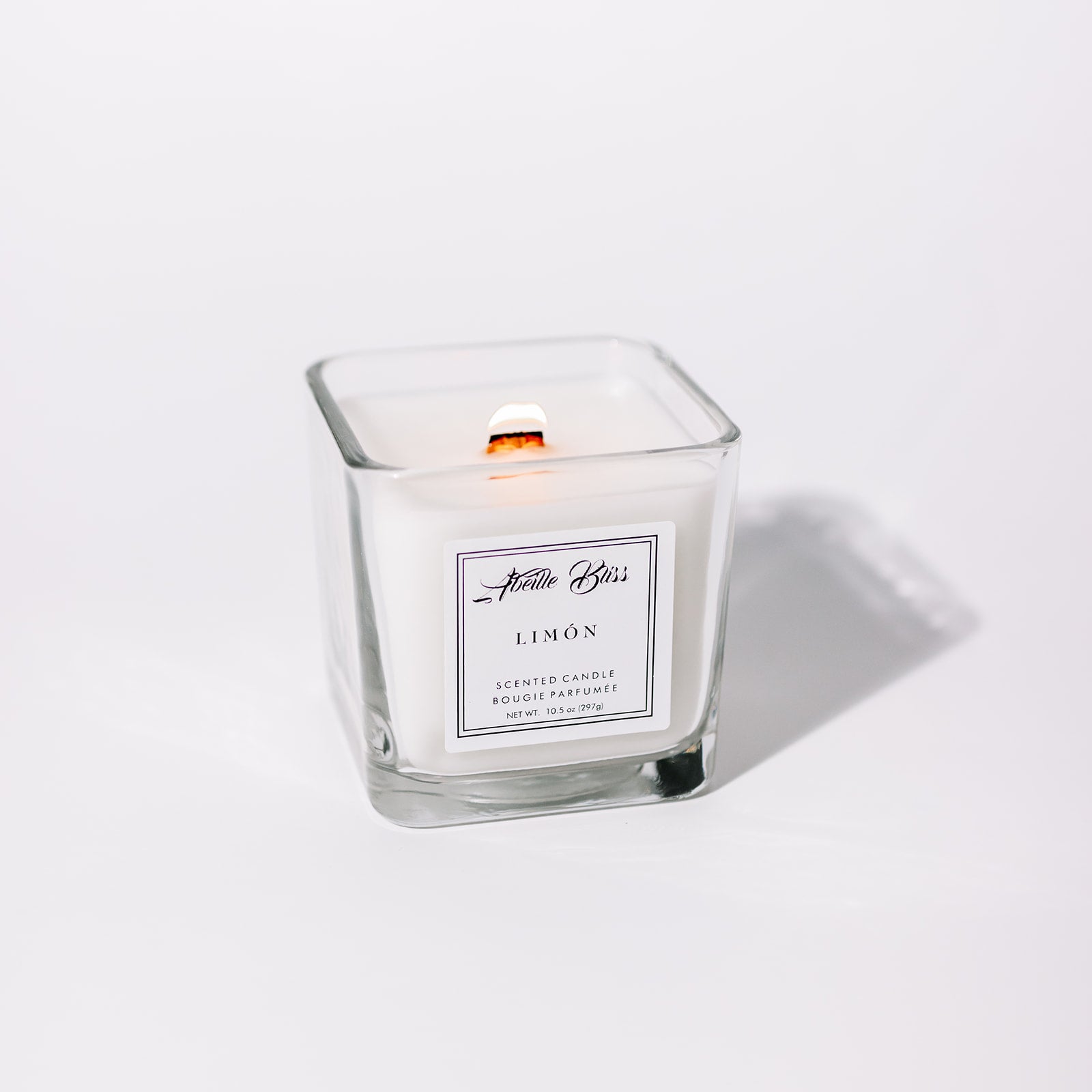 A white candle in a clear glass jar with a label that reads 'LIMÓN'. The candle is scented and has a wooden wick.