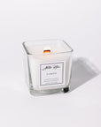 A white candle in a clear glass jar with a label that reads 'LIMÓN'. The candle is scented and has a wooden wick.