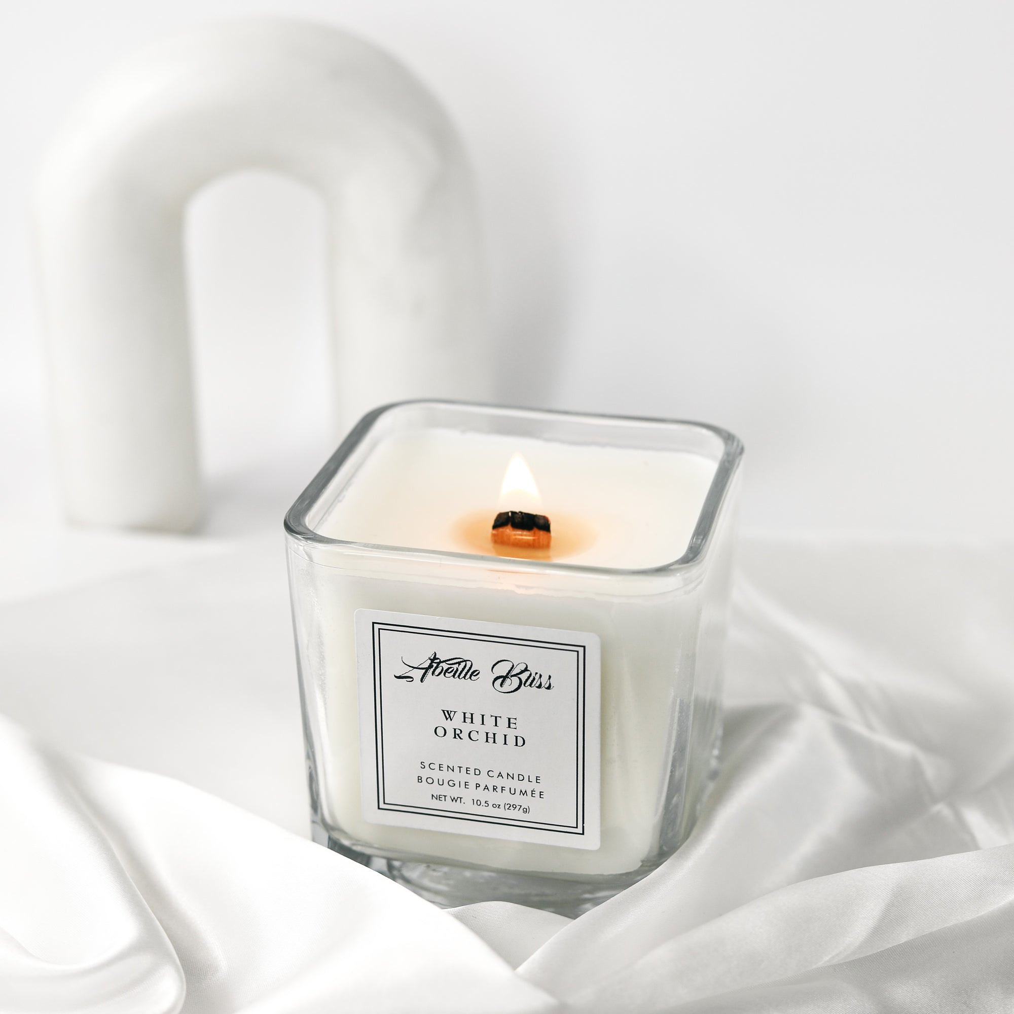 A white orchid scented candle with a label that reads 'White Orchid'. The candle is placed on a white, textured surface and has a wood wick.