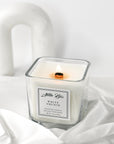 A white orchid scented candle with a label that reads 'White Orchid'. The candle is placed on a white, textured surface and has a wood wick.